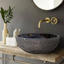 Toros Black Marble Above-Vanity Bathroom Vessel Sink Rough Exterior (D)16" (H)6" installed above wooden vanity with metal frames gold color faucet and toothbrushes and toothpaste bathroom set with beige color vase and green flowers
