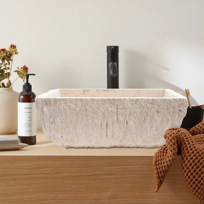 Troia Light Rustic Travertine Above Vanity Bathroom Sink Honed Interior Hand Chiseled Exterior installed bathroom above wooden vanity and black faucet brown towel on vanity tooth brush and liquid soap flower in vase