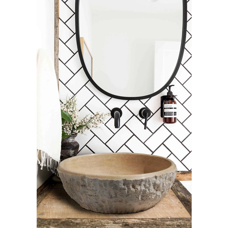 Troia Light Beige Travertine Above Vanity Bathroom Sink Dragon Split-face Exterior (D)16" (H)6" bathroom view installed above wooden vanity and white herringbone wall tiles and black wall-mount faucet hand-liquid soap mounted on wall and round black framed mirror wide view